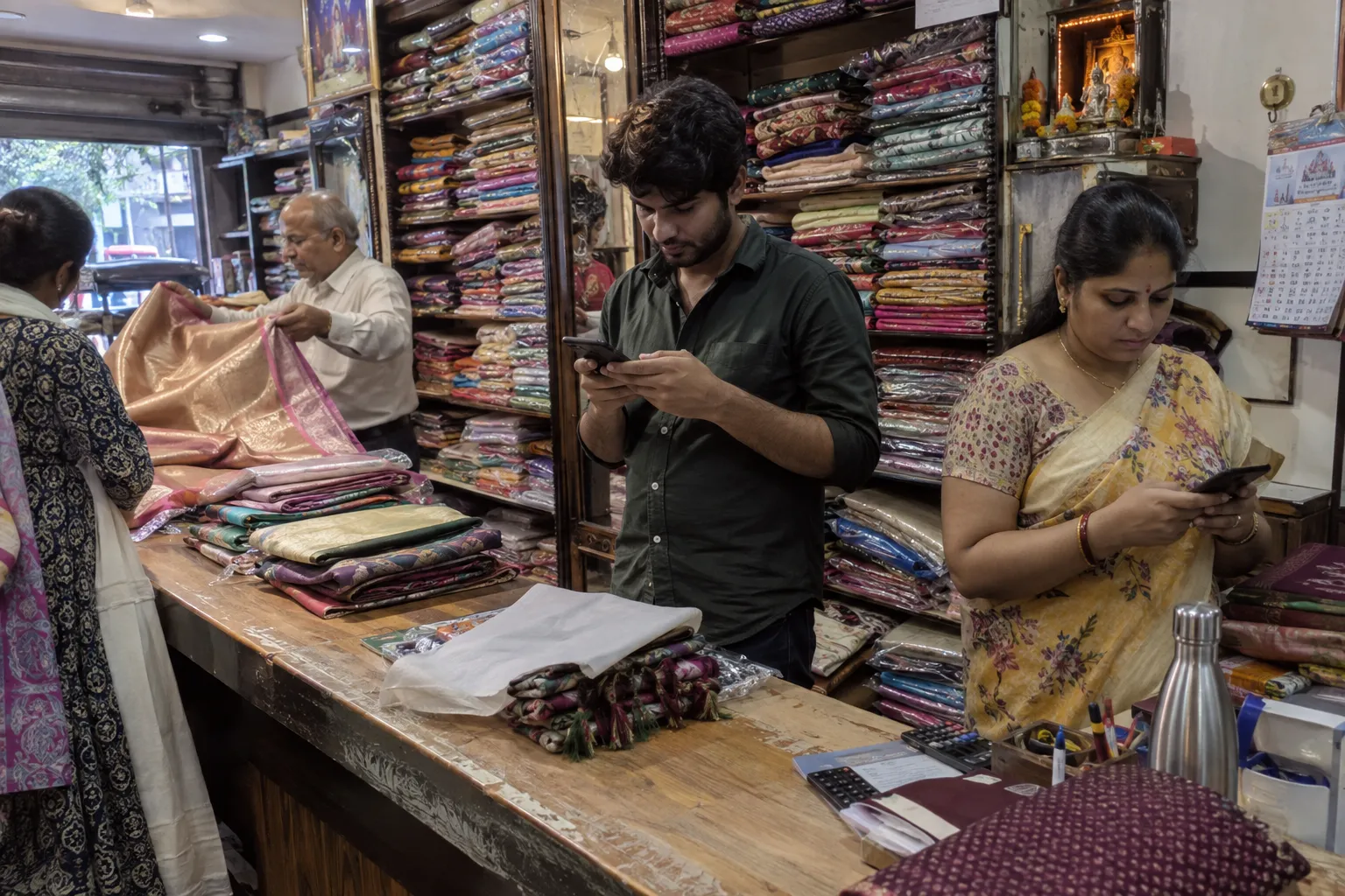 Two shop staff replying to customers on their phones while the owner shows a saree to a walk in customer.